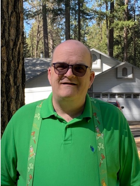 Image of a man wearing sunglasses, wearing a green polo like shirt with green suspenders, standing in front of a garage on a sunny day with redwood trees in the background.