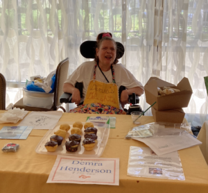 Image of Demra smiling and sitting behind a table with her baked cupcakes displayed.