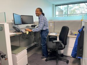 A man smiling and working at a standing desk inside of a cubicle in an office.