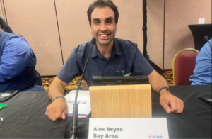 Councilmember Alex Reyes smiling and sitting down in front of a microphone attending a Council meeting.